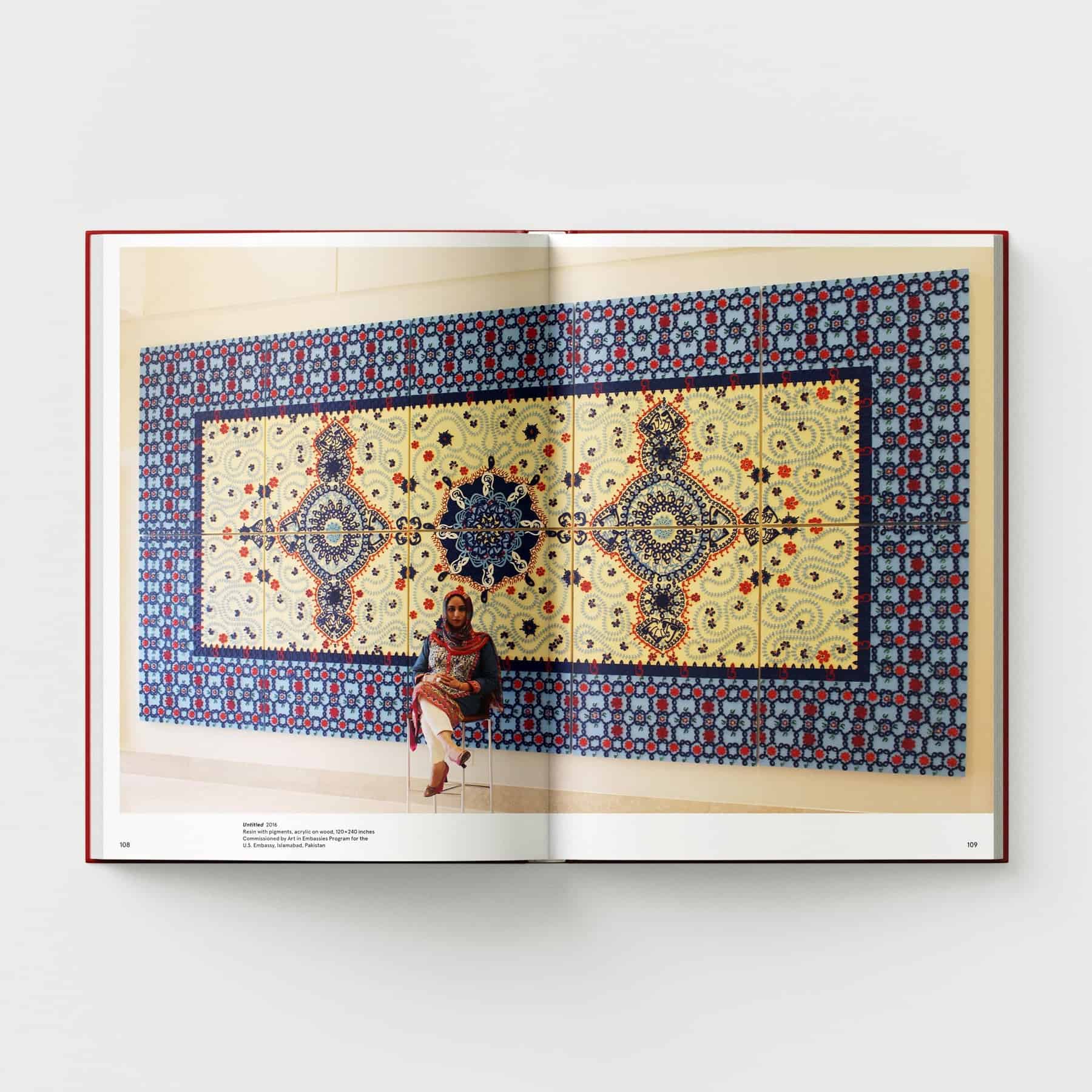 A woman sits before a page of "Ambreen Butt: What Comes to My Lips" by Black Dog Online, where a colorful tapestry with intricate patterns and geometric designs unfolds, reminiscent of an Indo-Persian miniature painting.