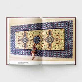 An open book titled "Ambreen Butt: What Comes to My Lips" from Black Dog Online showcases an artwork of a large, intricately patterned textile on a wall, reminiscent of Indo-Persian miniature painting. A person sits on the floor in front of the textile, gazing at its fusion with contemporary Western art elements.