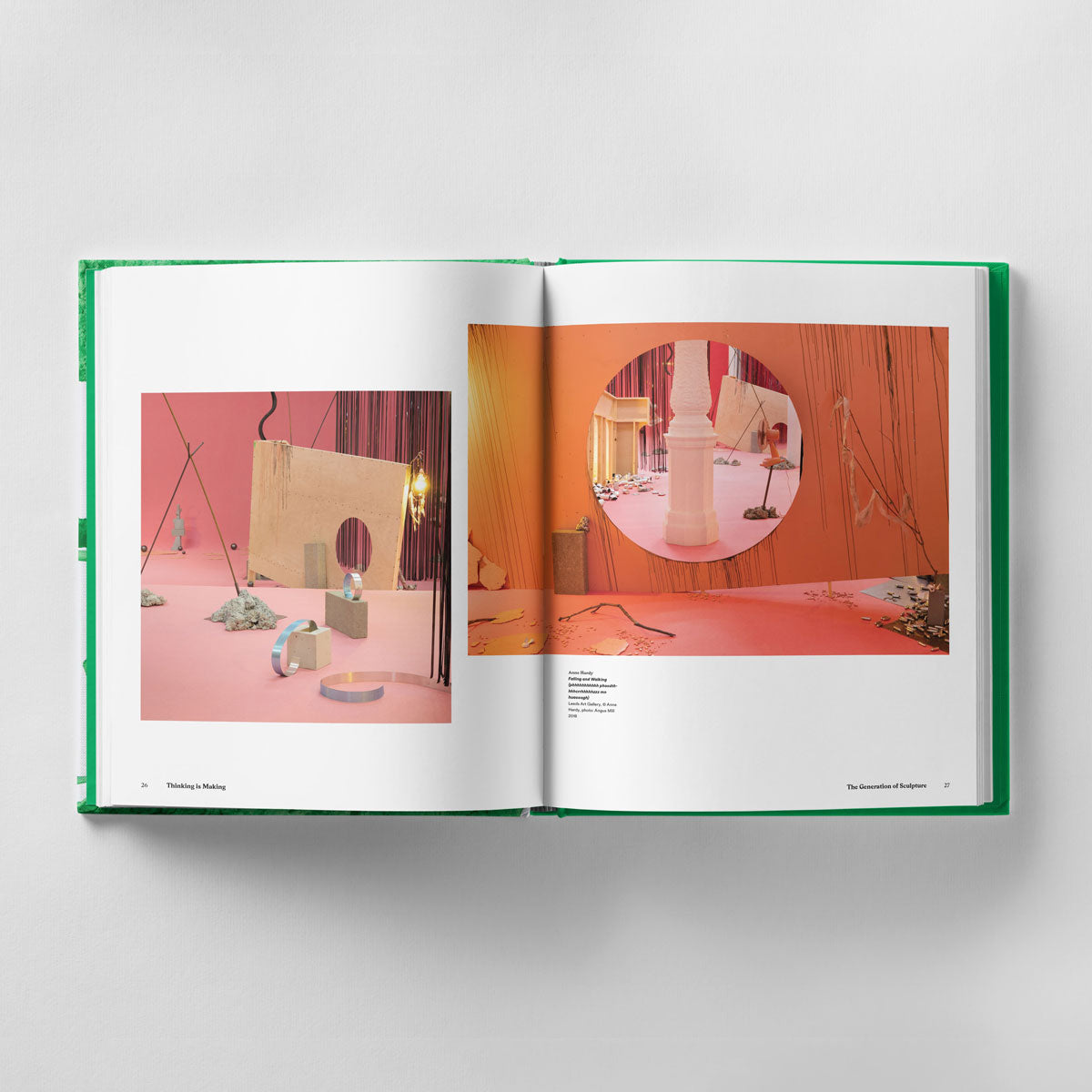 Open book displaying two pages of an artistic installation by emerging sculptors, featuring suspended objects, a peach-colored background, and circular focus frames. The book rests on a white surface with its green cover partially visible, showcasing **Thinking is Making: Objects in Space The Mark Tanner Sculpture Award by Black Dog Online**.