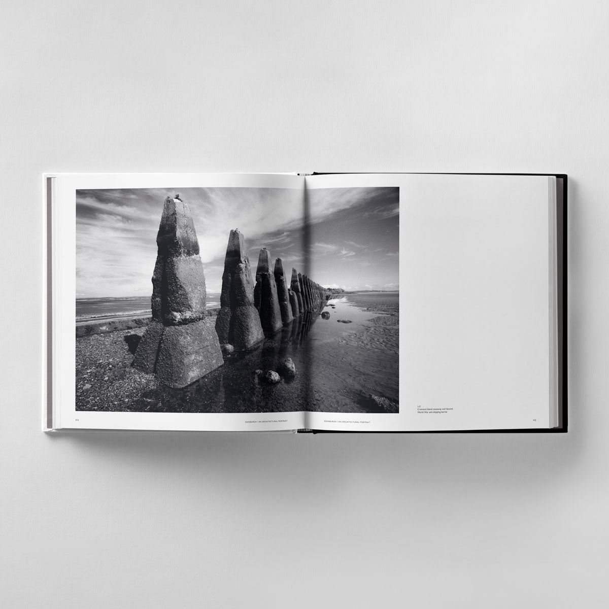 An open book showcases an evocative black-and-white photograph by James Reid, featuring weathered stones on a beach under a cloudy sky—a nod to architectural photography in "Edinburgh: An Architectural Portrait" by Black Dog Online.