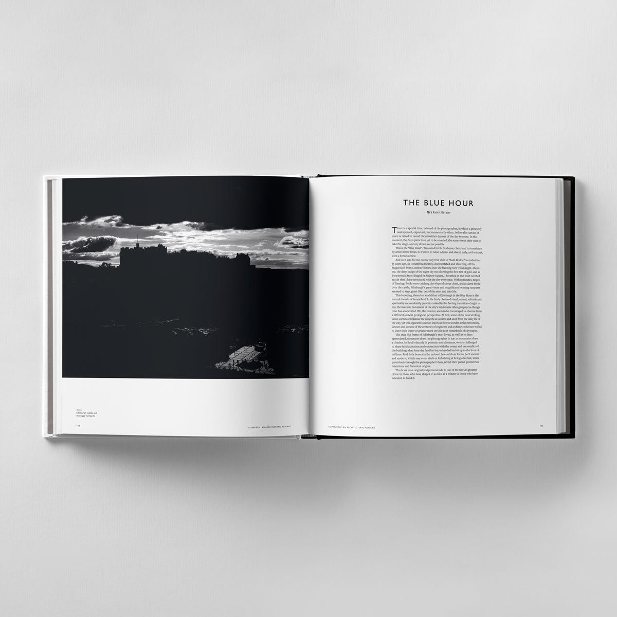 A book, "Edinburgh: An Architectural Portrait" by Black Dog Online, lies open on a gray surface; the left page displays a black and white photo of Edinburgh's architecture. The right page, titled "The Blue Hour," features text that complements the scene in James Reid's photographic style.