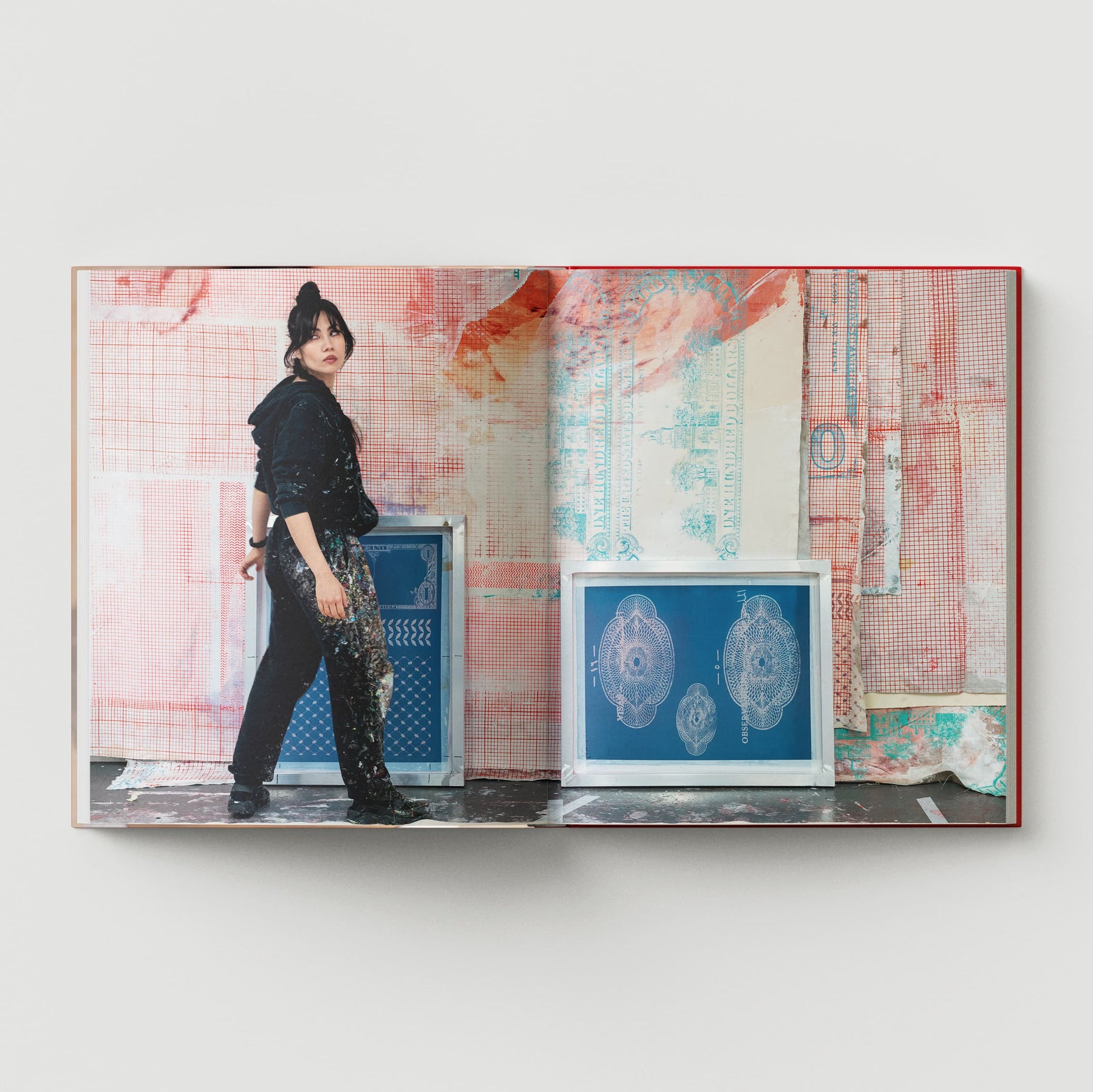 A person stands in front of a vibrant piece by contemporary artist Mandy El-Sayegh, clad in black attire. The backdrop showcases abstract patterns in red, blue, and white that echo the designs featured in "The Makeshift Body: Mandy El-Sayegh," a monograph by Black Dog Online.