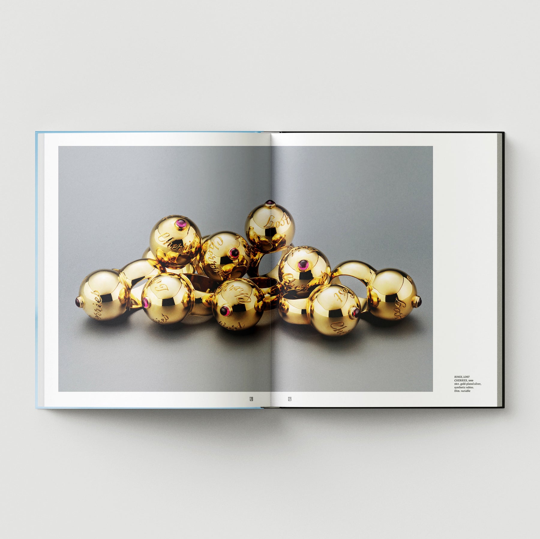 An open book from Black Dog Press, "Formula + Fetish," displays a Mark Woods photograph of gold spheres with red details on gray—a striking blend of jewellery and fine art.