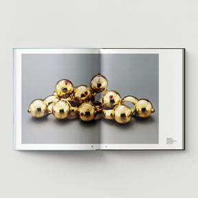 An open book from Black Dog Press, "Formula + Fetish," displays a Mark Woods photograph of gold spheres with red details on gray—a striking blend of jewellery and fine art.