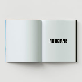 An open book from Black Dog Press titled "Formula + Fetish" displays a blank left page and a right page with "PHOTOGRAPHS" in bold, black letters—an evocative piece reminiscent of British artist Mark Woods' jewellery and fine art.