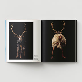 A book spread from "Formula + Fetish" by Black Dog Press showcases two photos by British artist Mark Woods, where people wear antlers and draped clothing against a black background, blending jewellery with fine art.