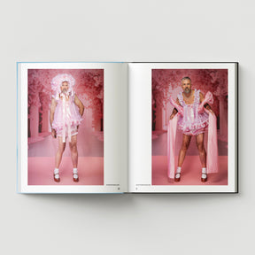 An open book from Black Dog Press’s "Formula + Fetish" shows two photos of a bearded man in a pink frilly dress, bonnet, and white socks against a pink background, reflecting Mark Woods’ fusion of jewellery and fine art.