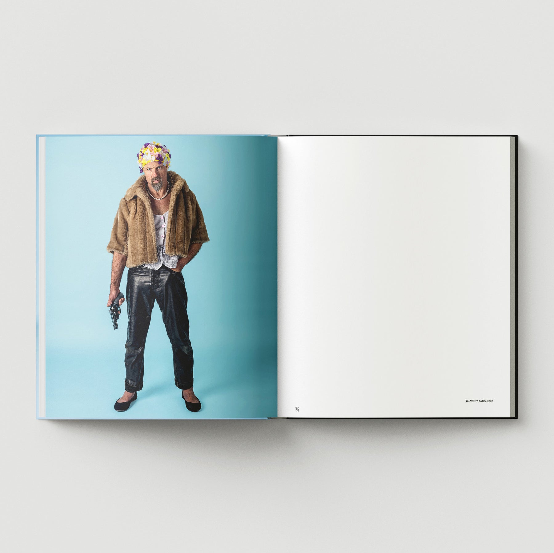 Someone stands on a blue background holding a handgun, dressed in eclectic style inspired by Mark Woods. They wear a faux fur jacket, black pants, flats, and a flowered headpiece. The facing page is blank. From "Formula + Fetish" by Black Dog Press.