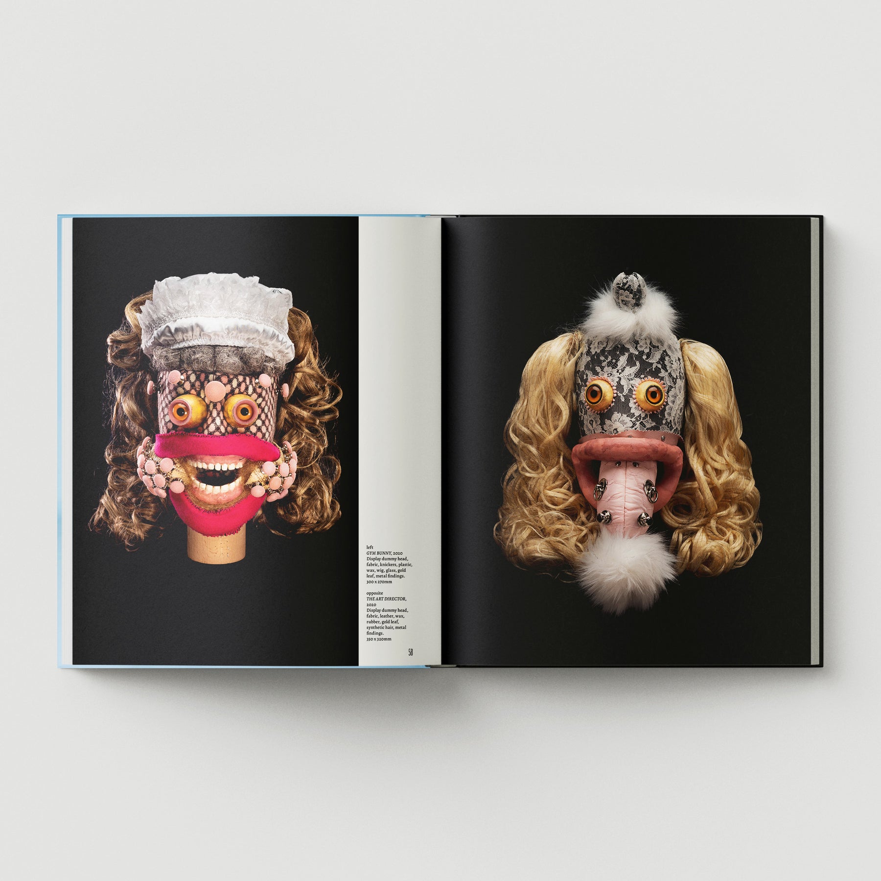 An open book titled "Formula + Fetish" by Black Dog Press displays photos of sculptural masks with exaggerated features and blonde wigs, set against black backgrounds, highlighting the unique blend of jewelry and fine art.